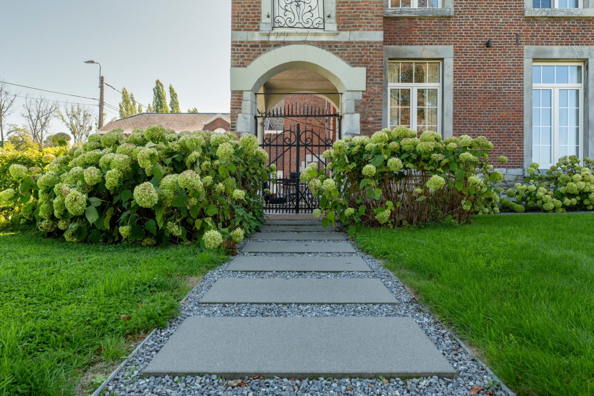 Allée en dalles sur gravier menant au portail d'un manoir, bordée d'hortensias