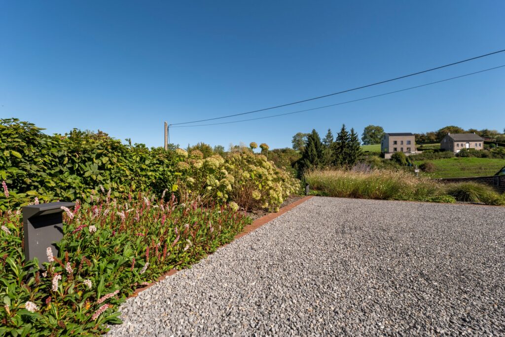 Cour en gravier bordée d'un massif de renouées et d'hortensias, avec vue sur la campagne