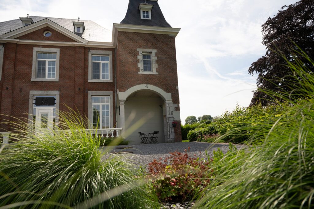 Façade d'un manoir en briques vue à travers des graminées, avec cour en gravier